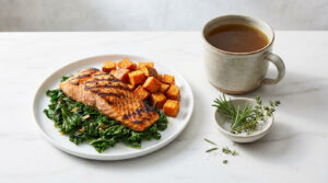 Grilled salmon fillet on kale with roasted sweet potatoes and a cup of broth beside fresh herbs on a plate.