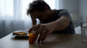 A man with his head in his hand reaches for a pill bottle on a table with toast and a glass of water nearby.