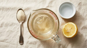A measuring cup with liquid, a spoon of sugar, a lemon half, and a small dish on a linen surface.