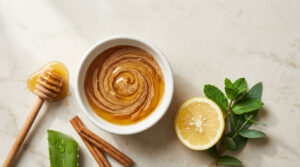 A bowl of honey with a swirl, lemon slice, aloe vera, mint leaves, and cinnamon sticks on a marble surface.