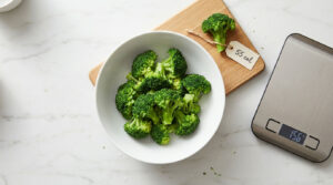 Fresh broccoli florets in a white bowl on a marble surface with a digital scale nearby.