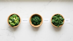 Three wooden bowls contain broccoli, spinach, and kale on a marble countertop with labels beside each bowl.