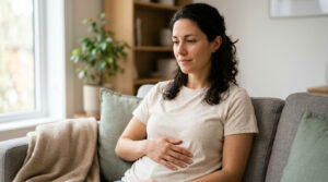 A woman sits on a couch, resting her hand on her stomach, with a plant and soft furnishings in the background.