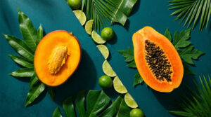 A halved papaya and a whole papaya on green leaves with lime slices and palm fronds in a vibrant arrangement.