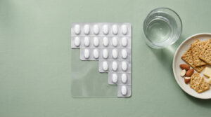 A blister pack of white pills sits next to a glass of water and a plate of crackers, cheese, and nuts on a green background.
