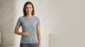 A woman in a gray t-shirt stands with her hand on her stomach, featuring a subtle illustration on the shirt.