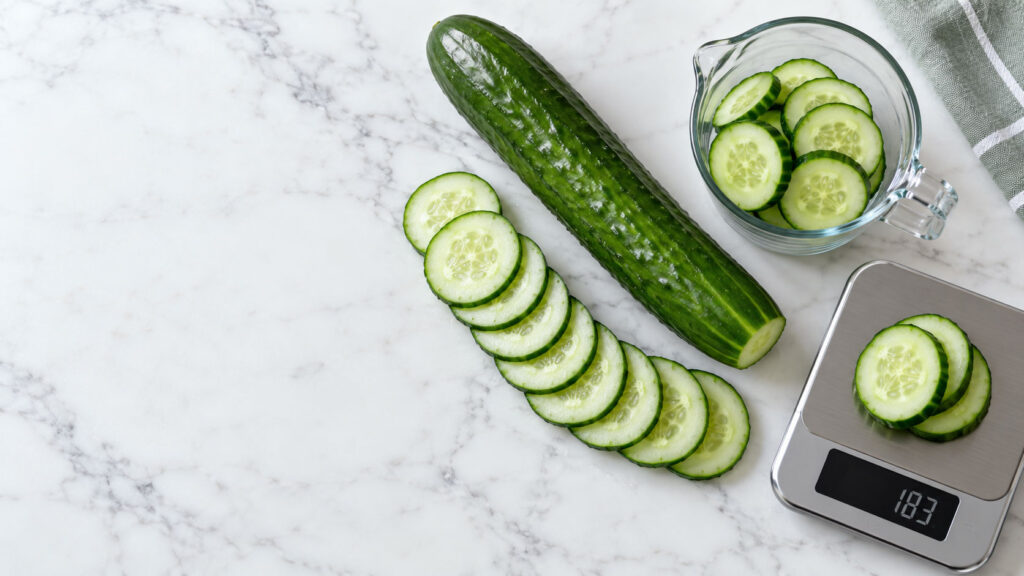 Fresh cucumber slices arranged on a marble surface with a whole cucumber and a measuring scale nearby.
