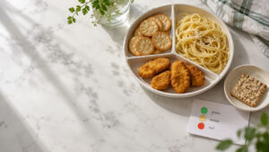 A plate with chicken nuggets, crackers, spaghetti, and a snack on a marble table with a plant nearby.