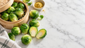 Fresh Brussels sprouts in a bamboo steamer with cut pieces and a bowl of seasoning on marble surface.