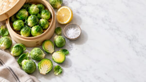 Fresh Brussels sprouts in a bamboo steamer with lemon and salt on a marble surface.