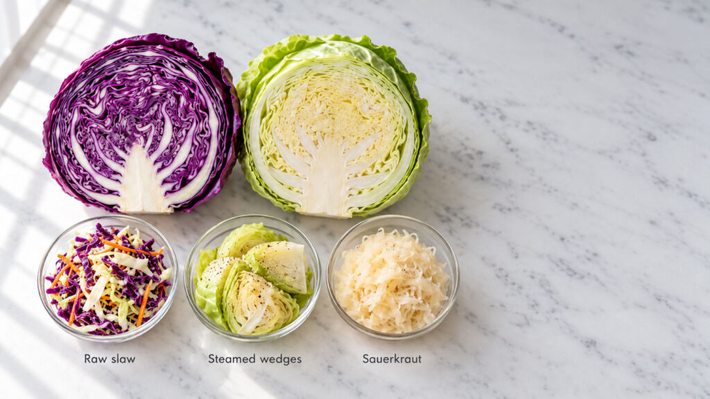 A purple cabbage half, a green cabbage half, and bowls of raw slaw, steamed wedges, and sauerkraut on a marble surface.