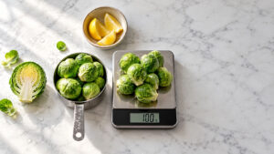 Fresh Brussels sprouts and sliced cabbage on a marble countertop with a digital scale showing 100 grams.