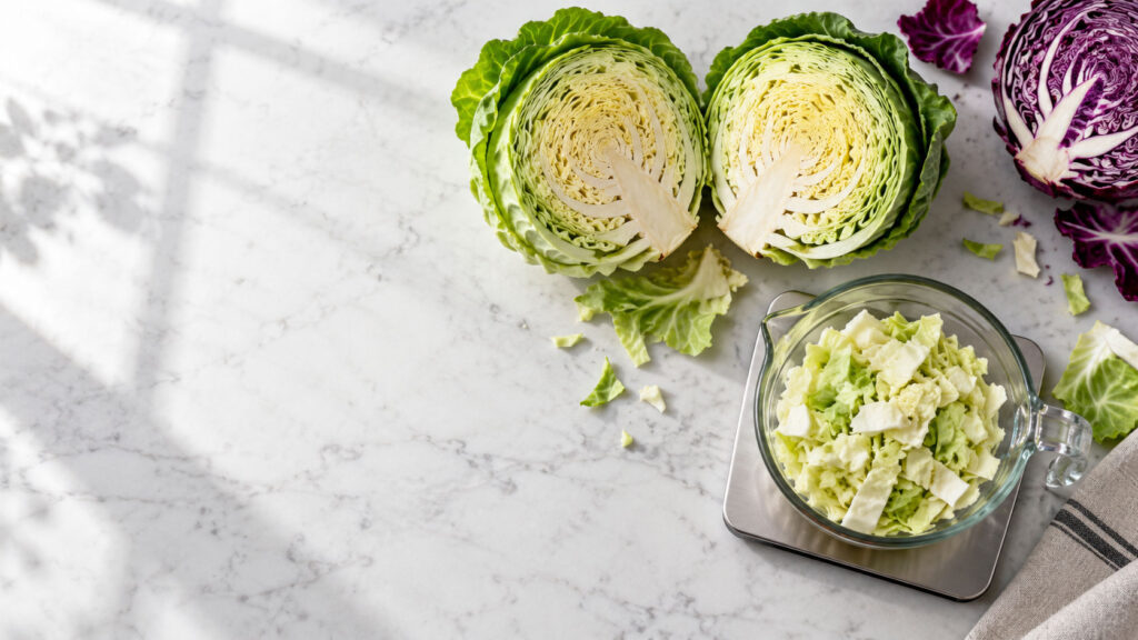 Freshly chopped green and purple cabbage on a marble countertop with shadows and a measuring cup.