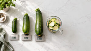 Three zucchinis of varying sizes on digital scales with sliced zucchini in a measuring cup nearby.