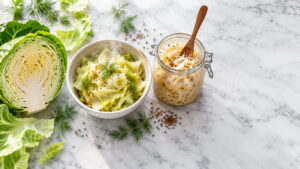 Fresh green cabbage, a bowl of cooked cabbage, and a jar of sauerkraut on a marble surface.